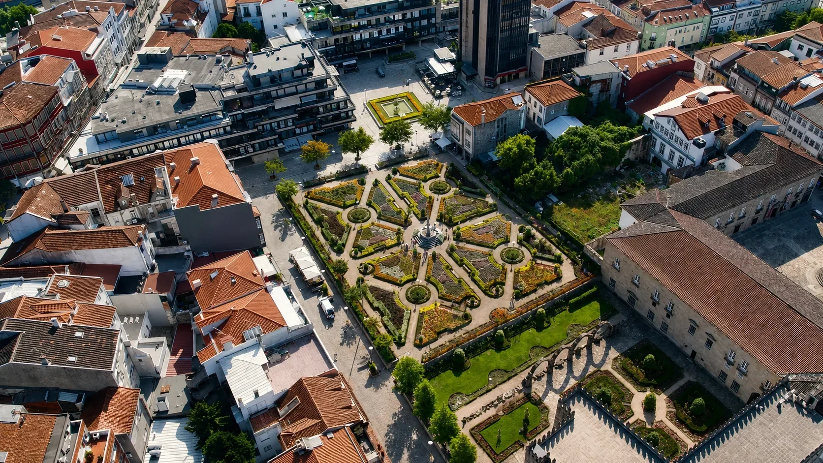 The Santa Bárbara Garden in Braga — one of Portugal's most photographed baroque gardens, showing the formal parterre with clipped box hedges, central fountain, and seasonal flowers set against the dramatic backdrop of the 14th-century tower of the Archbishops' Palace (Paço dos Arcebispos), a composition beloved by artists and photographers for centuries