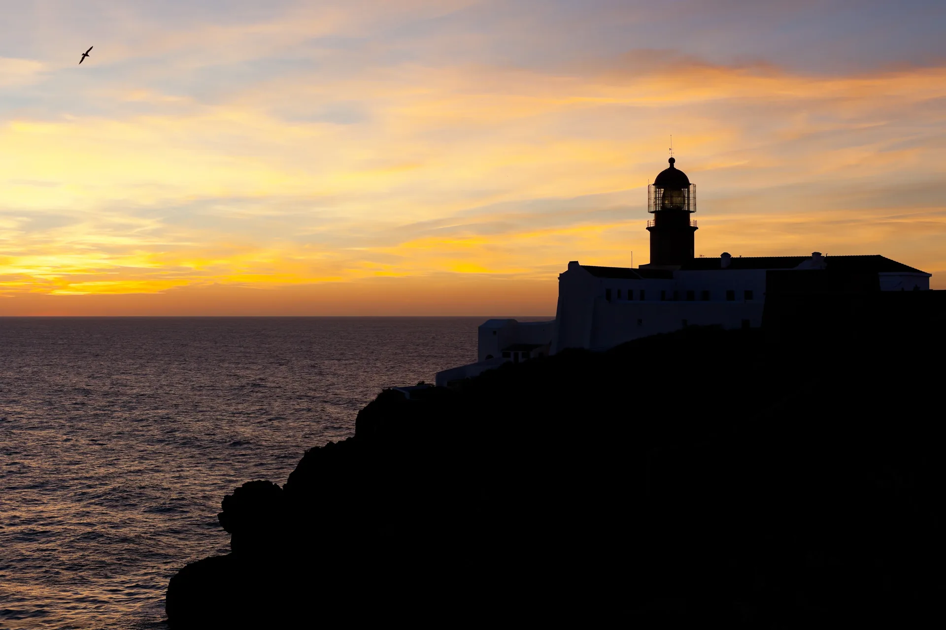 Cape St. Vincent bathed in late afternoon golden light — the dramatic headland and lighthouse at the far southwest corner of continental Europe with the Atlantic Ocean glowing in warm orange and amber tones as the sun descends towards the horizon